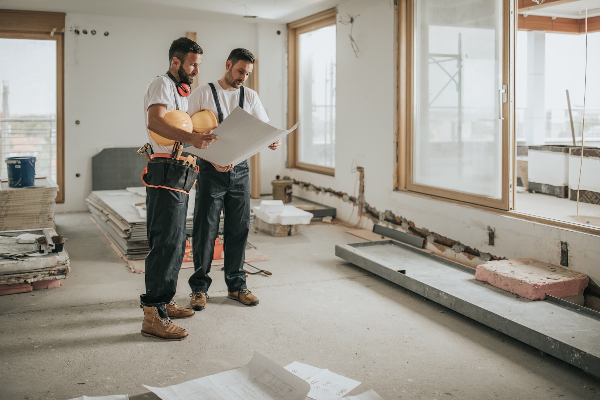 Two construction workers reviewing plans inside a building amid stacks of drywall.